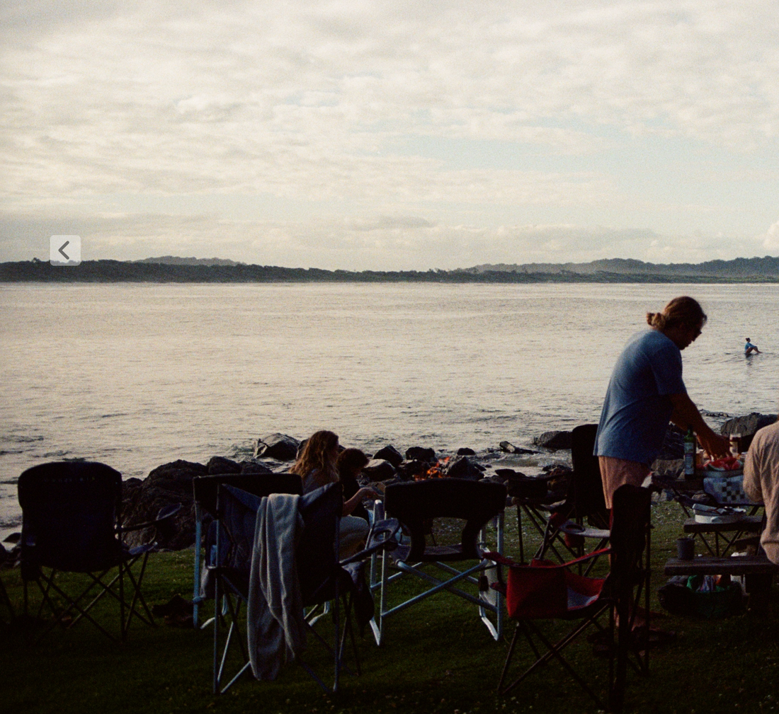 A group of dads around a fire on a rocky beach at sunset, one cooking at a table by the water
