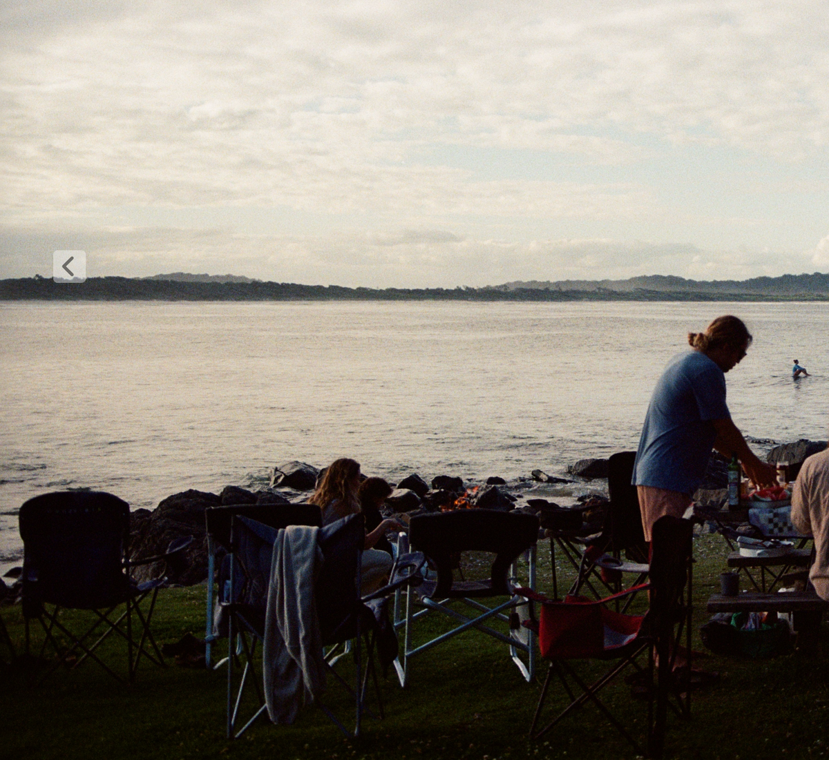 A group of dads around a fire on a rocky beach at sunset, one cooking at a table by the water