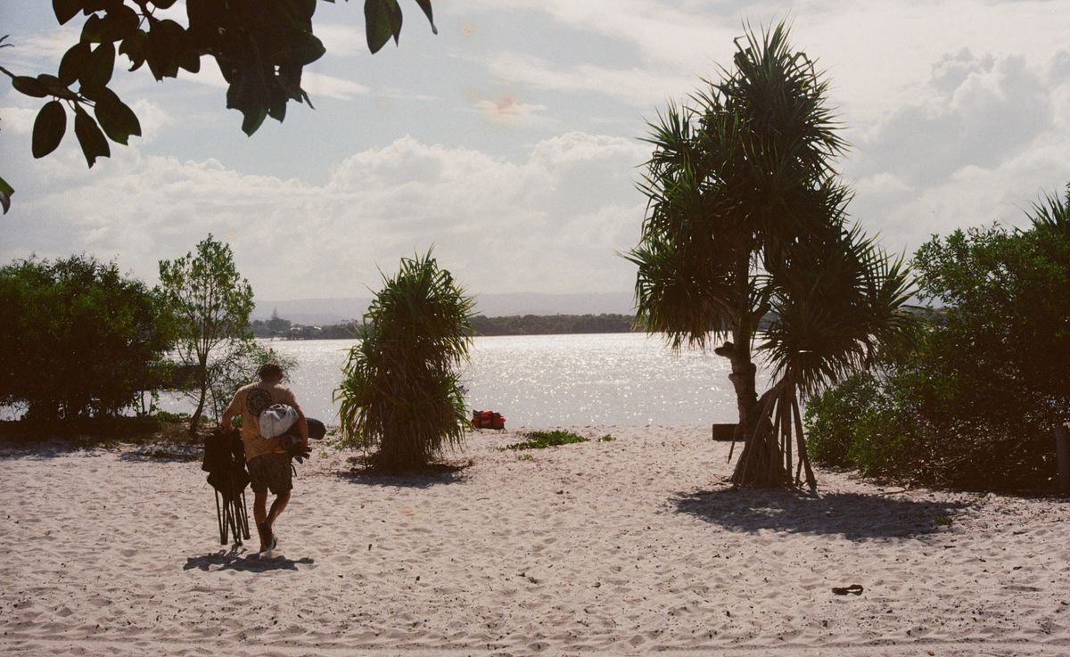 A dad carrying camp chairs and gear across a sandy beach toward a calm lake at dusk, pandanus palms around